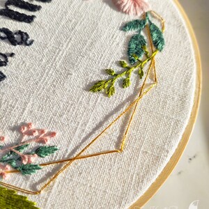 May include: A white embroidery hoop with a gold geometric design and floral embroidery. The embroidery includes pink and green flowers and leaves.