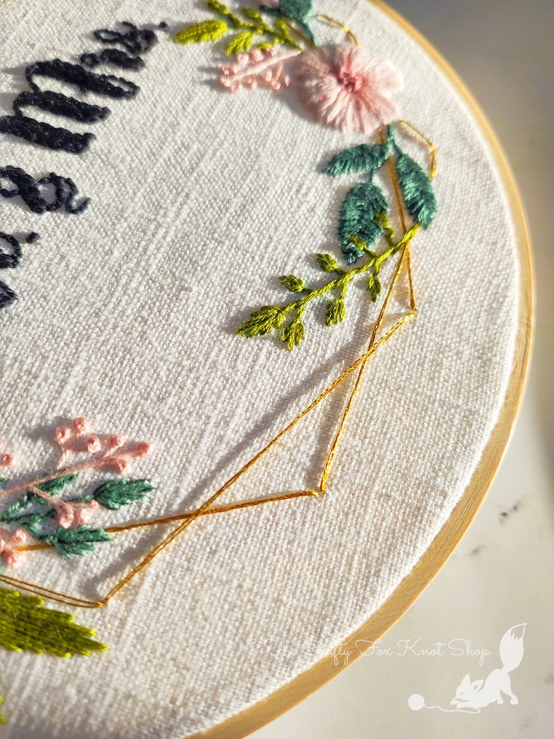 May include: A white embroidery hoop with a gold geometric design and floral embroidery. The embroidery includes pink and green flowers and leaves.