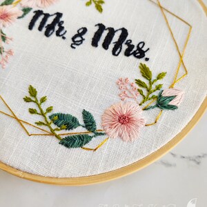 May include: Embroidered hoop art with a gold geometric frame and pink flowers. The text "Mr. & Mrs." is stitched in black thread.