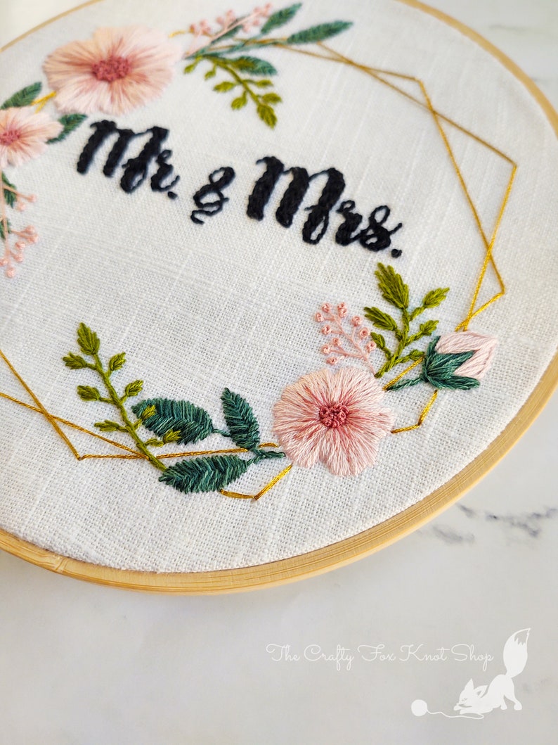 May include: Embroidered hoop art with a gold geometric frame and pink flowers. The text "Mr. & Mrs." is stitched in black thread.