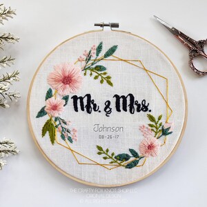 May include: Embroidered hoop art with a gold geometric frame and pink and green floral designs. The text "Mr. & Mrs. Johnson 08-26-17" is embroidered in black thread.