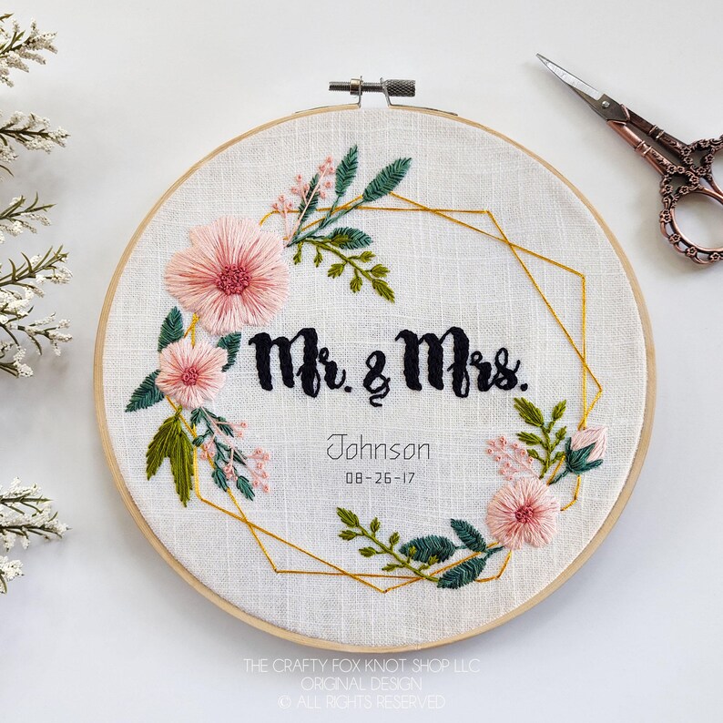 May include: Embroidered hoop art with a gold geometric frame and pink and green floral designs. The text "Mr. & Mrs. Johnson 08-26-17" is embroidered in black thread.