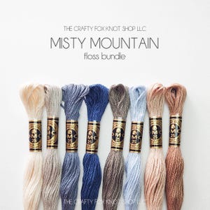 May include: A set of eight embroidery floss spools in various shades of beige, blue, and brown. The spools are labeled "DMC" and "25". The text "Misty Mountain floss bundle" is displayed above the spools.