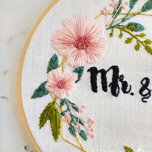 May include: Embroidered hoop art with pink flowers, green leaves, and gold geometric shapes. The text "Mr. &" is stitched in black thread.