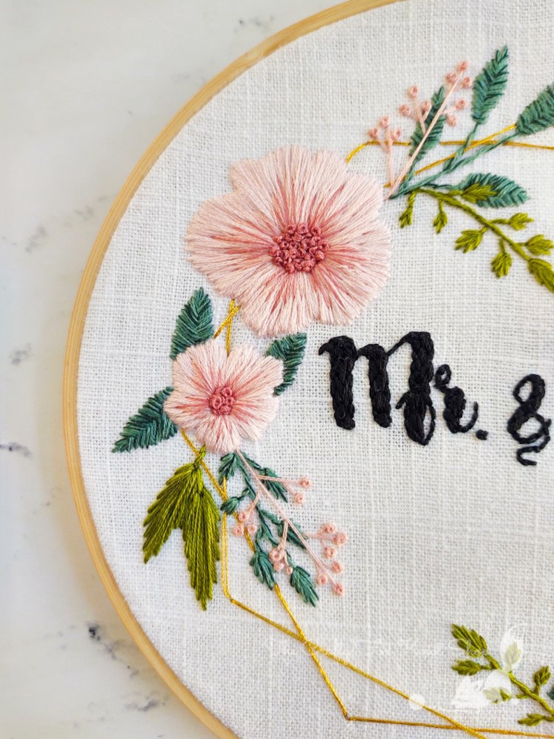 May include: Embroidered hoop art with pink flowers, green leaves, and gold geometric shapes. The text "Mr. &" is stitched in black thread.