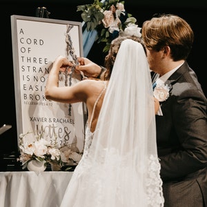 May include: A wedding ceremony with a sign that reads "A Cord of Three Strands Is Not Easily Broken." The sign includes the names Xavier & Rachael and the date 23-22. A bride and groom are braiding cords together. Flowers and candles are also visible.