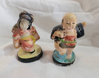 May include: Two vintage Japanese figurines, one depicting a woman in a pink and yellow kimono and the other a man in a blue and black robe. Both figurines are painted with intricate details and stand on small circular bases.