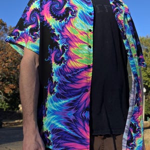 Op de afbeelding: Korte mouwen overhemd met een levendig, wervelend fractaalpatroon in paars, blauw, groen en geel op een zwarte achtergrond. Het overhemd is open, waardoor een zwart t-shirt zichtbaar is. Het heeft een klassieke kraag en een knoopsluiting.