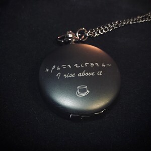 May include: A black pocket watch with a silver chain. The watch face has an engraved quote: "& p h = 3 2 1 6 9 1 4 ~ I rise above it".