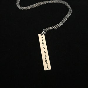 May include: A silver chain necklace with a silver pendant. The pendant is a rectangular bar with a vertical inscription of black symbols.