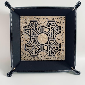 May include: A black leather dice tray with a gold interior featuring a black and white geometric design. The design includes a central circle and a repeating pattern of lines and shapes.