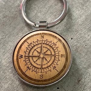 May include: A wooden compass keychain with a silver ring. The compass features a detailed design with the cardinal directions N, S, E, and W.