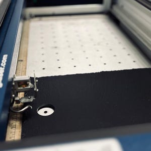 May include: A close-up of a laser cutter with a black material being cut. The material is on a white mat with a grid pattern. The laser cutter has a blue and silver exterior.