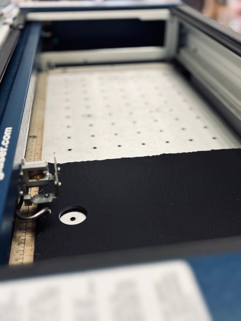 May include: A close-up of a laser cutter with a black material being cut. The material is on a white mat with a grid pattern. The laser cutter has a blue and silver exterior.
