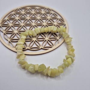 Peut inclure: Un bracelet en pierres vertes claires. Le bracelet est composé de petites pierres de forme irrégulière. Le bracelet repose sur un dessous de verre en bois avec un motif géométrique. Le fond est blanc.