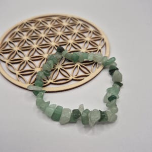 Peut inclure: Un bracelet de perles en éclats d'aventurine verte. Le bracelet est composé de petites pierres vertes de forme irrégulière. Le bracelet repose sur un dessous de verre en bois avec un motif géométrique de fleur de vie. Le fond est blanc.