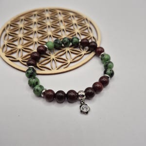 May include: A bracelet with maroon and green speckled beads, interspersed with silver-coloured spacers. A small, silver-coloured charm with a clear stone in a flower shape hangs from the bracelet. The bracelet is displayed on a wooden coaster.