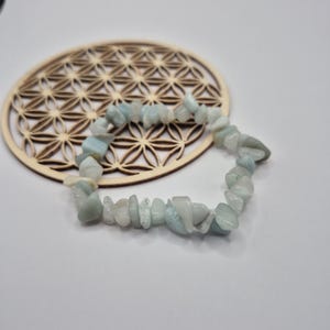 Peut inclure: Un bracelet de pierres précieuses bleu clair et blanches. Le bracelet est composé de petites pierres de forme irrégulière. Il repose sur un dessous de verre en bois avec un motif floral géométrique.