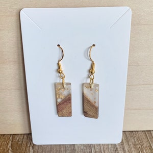 May include: A pair of gold dangle earrings with rectangular resin charms. The charms are translucent and contain wood and gold flakes.