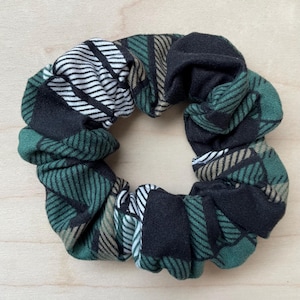 May include: A green, black, and white plaid scrunchie. The fabric is patterned with stripes and checks in shades of green, black, white, and tan. The scrunchie is circular and appears to be made of a soft fabric, suitable for hair styling.