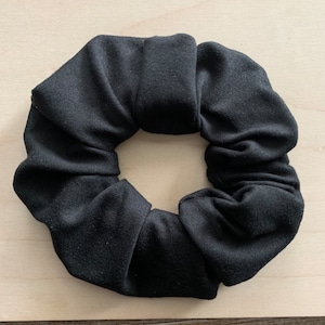 May include: A black scrunchie made from soft fabric. The hair accessory has a circular shape with a gathered design. The scrunchie is a solid black colour and is on a light wood surface.