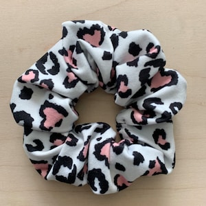 May include: A white scrunchie with a black and pink leopard print pattern. The fabric is gathered and elasticized, creating a voluminous, circular shape. The scrunchie is designed for use in hair styling.