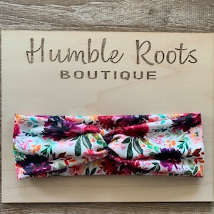 May include: A white headband with a floral print in shades of pink, purple, and green. The headband is twisted and has a knot in the center. The headband is displayed on a wooden surface with the text "Humble Roots Boutique" printed on it.