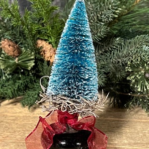 May include: A small blue bottle brush Christmas tree with a white flocked finish, decorated with a brown twig nest and a red ribbon bow.