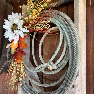 May include: A rustic wreath made with a coiled rope and decorated with dried flowers, wheat, and berries. The wreath is hanging on a weathered wooden wall.