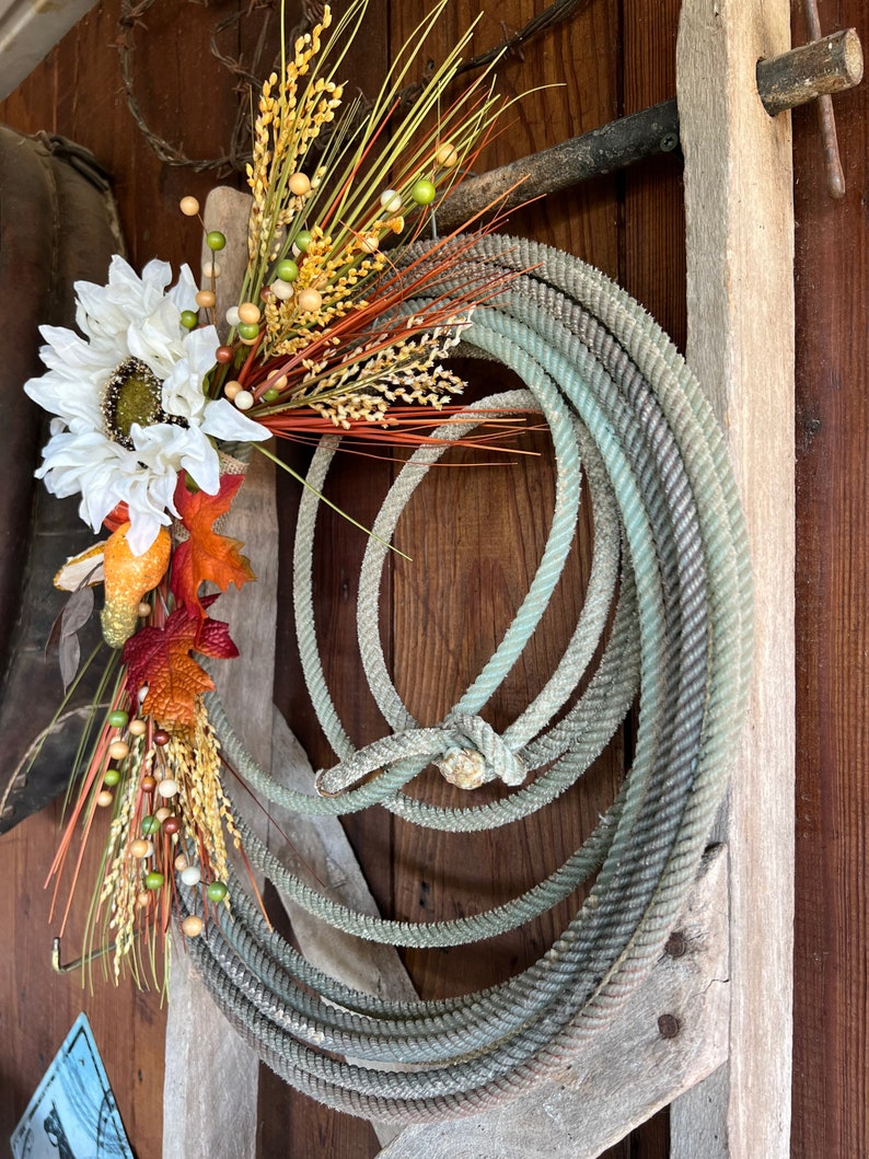 May include: A rustic wreath made with a coiled rope and decorated with dried flowers, wheat, and berries. The wreath is hanging on a weathered wooden wall.