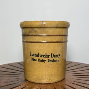 May include: A tan-colored, cylindrical stoneware crock with the text "Landwehr Dairy Fine Dairy Products" in black lettering. The crock has a slightly glossy finish and horizontal bands around the body.