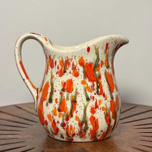 May include: A ceramic pitcher with a cream-colored base, adorned with vibrant orange and dark green splatters. The pitcher has a curved handle and a spout for easy pouring. The speckled design adds a unique, artistic touch to this kitchenware item.