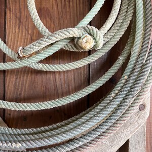 May include: A close-up of a coiled rope made of thick, braided, light green cord. The rope is wrapped around itself and tied in a knot. The rope is resting on a weathered wooden surface.