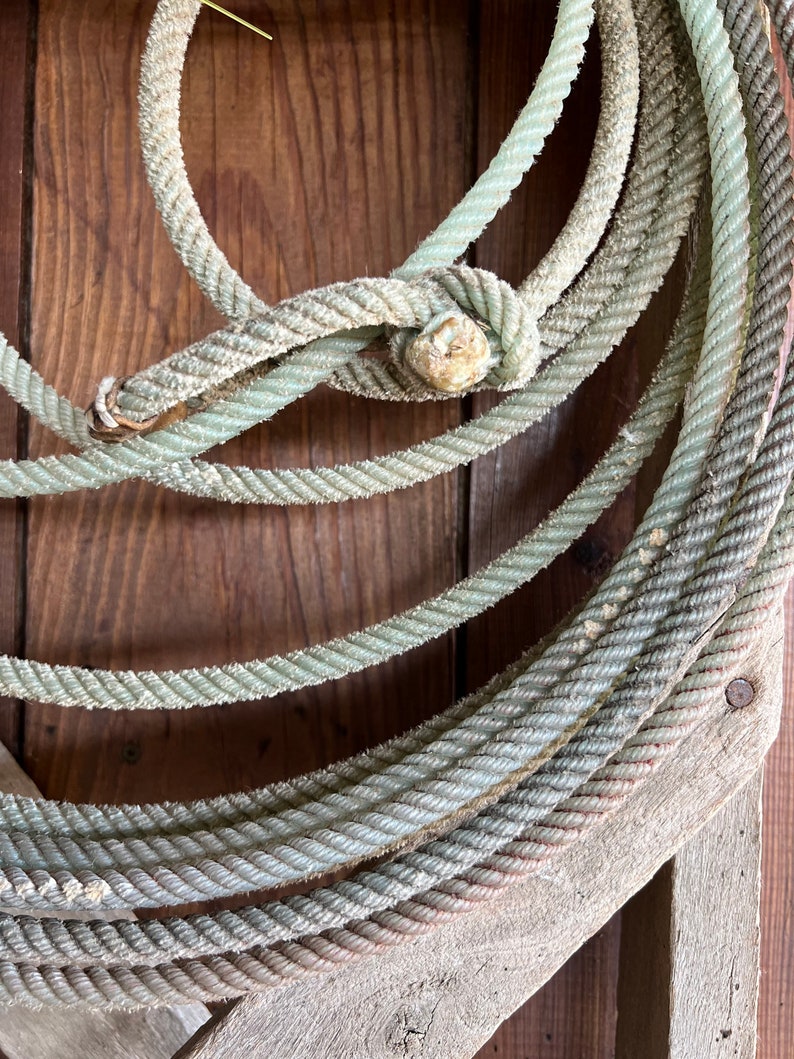 May include: A close-up of a coiled rope made of thick, braided, light green cord. The rope is wrapped around itself and tied in a knot. The rope is resting on a weathered wooden surface.