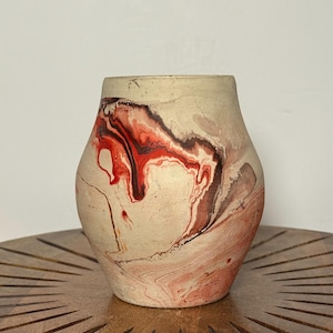 May include: A beige ceramic vase with a marbled design in shades of red, brown, and cream. The vase has a rounded body and a slightly flared rim. The decorative vase is displayed on a wooden table.