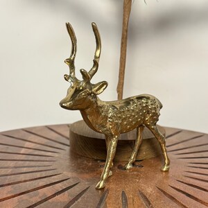May include: A small, gold-colored deer figurine with detailed antlers and textured body. The deer is standing on a wooden surface. The figurine is a decorative item, likely for home decor.