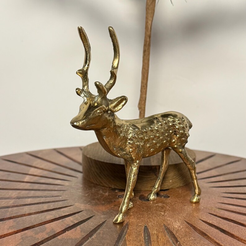 May include: A small, gold-colored deer figurine with detailed antlers and textured body. The deer is standing on a wooden surface. The figurine is a decorative item, likely for home decor.