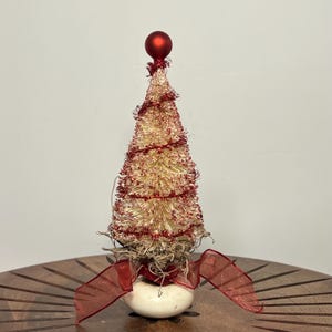 May include: A small, decorative Christmas tree with a gold and red color scheme. The tree is adorned with red tinsel and topped with a red ball. It sits on a white base with a red ribbon and decorative accents.
