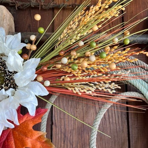 May include: A fall-themed wreath with a white sunflower, orange maple leaf, and brown, green, and beige wheat stalks with wooden beads.