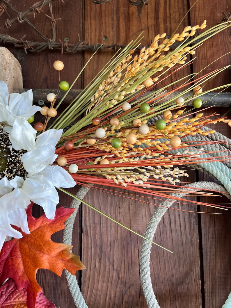 May include: A fall-themed wreath with a white sunflower, orange maple leaf, and brown, green, and beige wheat stalks with wooden beads.