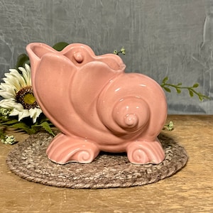 May include: A pink ceramic seashell planter with a detailed spiral design. The planter has three feet and is perfect for small plants or succulents.