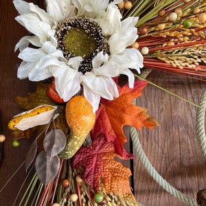 May include: A fall-themed wreath with a white faux sunflower, brown gourds, red and orange leaves, and wheat stalks with brown and green beads.
