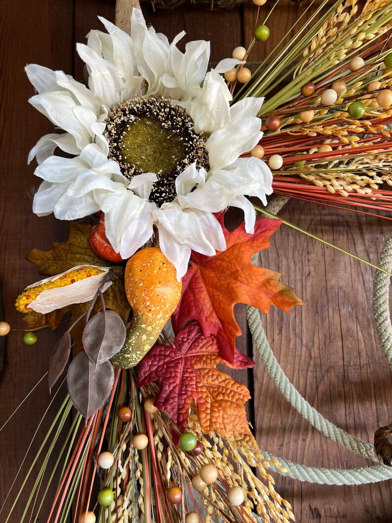 May include: A fall-themed wreath with a white faux sunflower, brown gourds, red and orange leaves, and wheat stalks with brown and green beads.