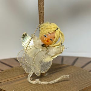 May include: A vintage Christmas angel ornament. The angel has a plastic face with blue eyes and yellow yarn hair. It wears a cream-colored dress with gold wings and a gold halo. The ornament is attached to a wooden dowel.