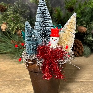 May include: A small wooden pot with a snowman figurine and three miniature flocked Christmas trees. The trees are white, green, and beige. The pot is decorated with red tinsel.