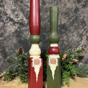 May include: Two wooden candle holders painted to look like Santa Claus. The candle holders are red and green with white accents. The Santa Claus faces are painted on the front of the candle holders.