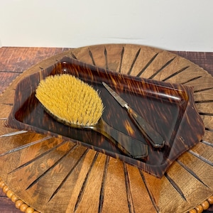 May include: A vintage vanity set featuring a tortoiseshell-patterned tray, a hairbrush with yellow bristles, and a small knife. The tray is rectangular with rounded corners. The set is displayed on a wooden decorative piece.