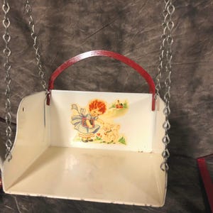 May include: A white metal swing set with a red metal frame and a picture of a girl and a lamb. The swing set is hanging from two silver chains.