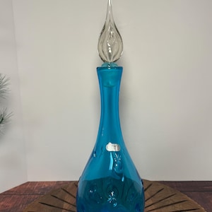 May include: A tall, turquoise glass bottle with a clear, teardrop-shaped stopper. The bottle has a slender neck and a bulbous base with decorative indentations. A small label is affixed to the side.
