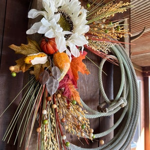 May include: A rustic fall wreath made with a coiled rope, white faux sunflowers, dried grasses, and small orange and brown gourds.